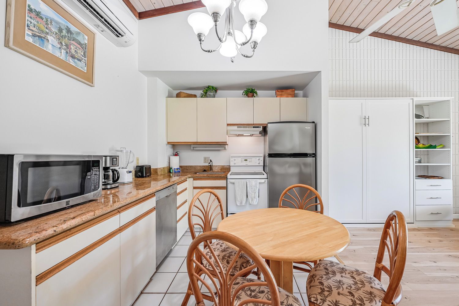 A compact kitchen with a microwave, toaster, and refrigerator. There is a dining table with chairs and a chandelier above.