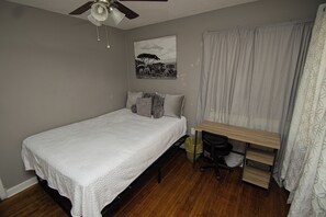 A bedroom with a bed, a desk, a chair, a ceiling fan, a window with curtains, and a wall-mounted picture.