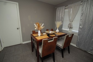 A dining area with a table set for four, chairs, a vase with dried plants, and a window with curtains.