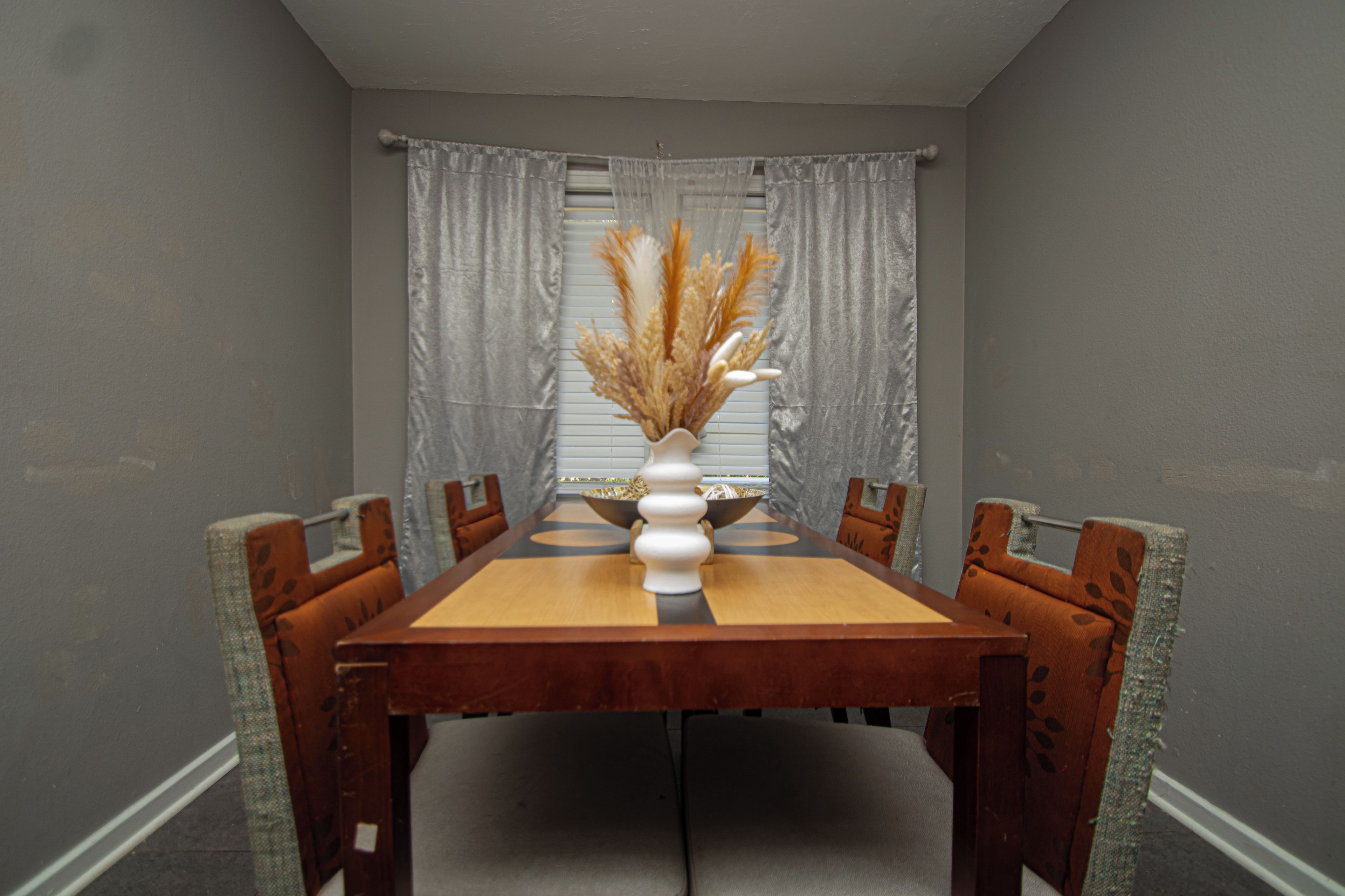 Une salle à manger avec une table en bois, six chaises, un vase contenant des plantes séchées et des rideaux gris.