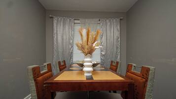 Une salle à manger avec une table en bois, six chaises, un vase contenant des plantes séchées et des rideaux gris.