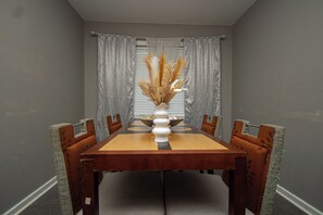 A dining room with a wooden table, six chairs, a vase with dried plants, and grey curtains.