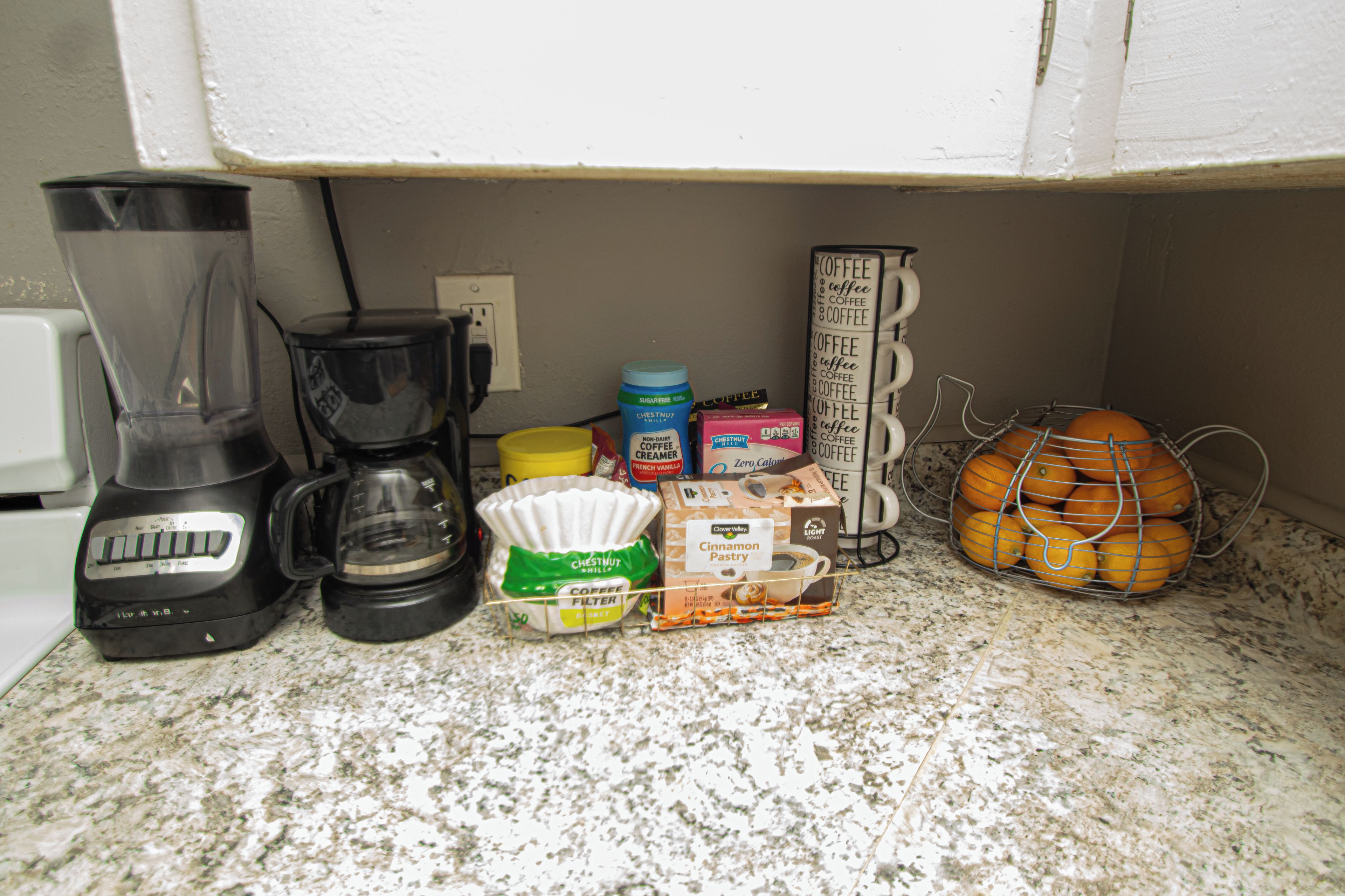 A kitchen counter with a coffee maker, a blender, a coffee filter basket, a jar of sugar, a box of creamer, and a basket of oranges.