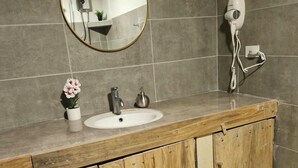 Ein Badezimmer mit einem Holztischchen, einem runden Spiegel, einem weißen Waschbecken und einem an der Wand montierten Fön.