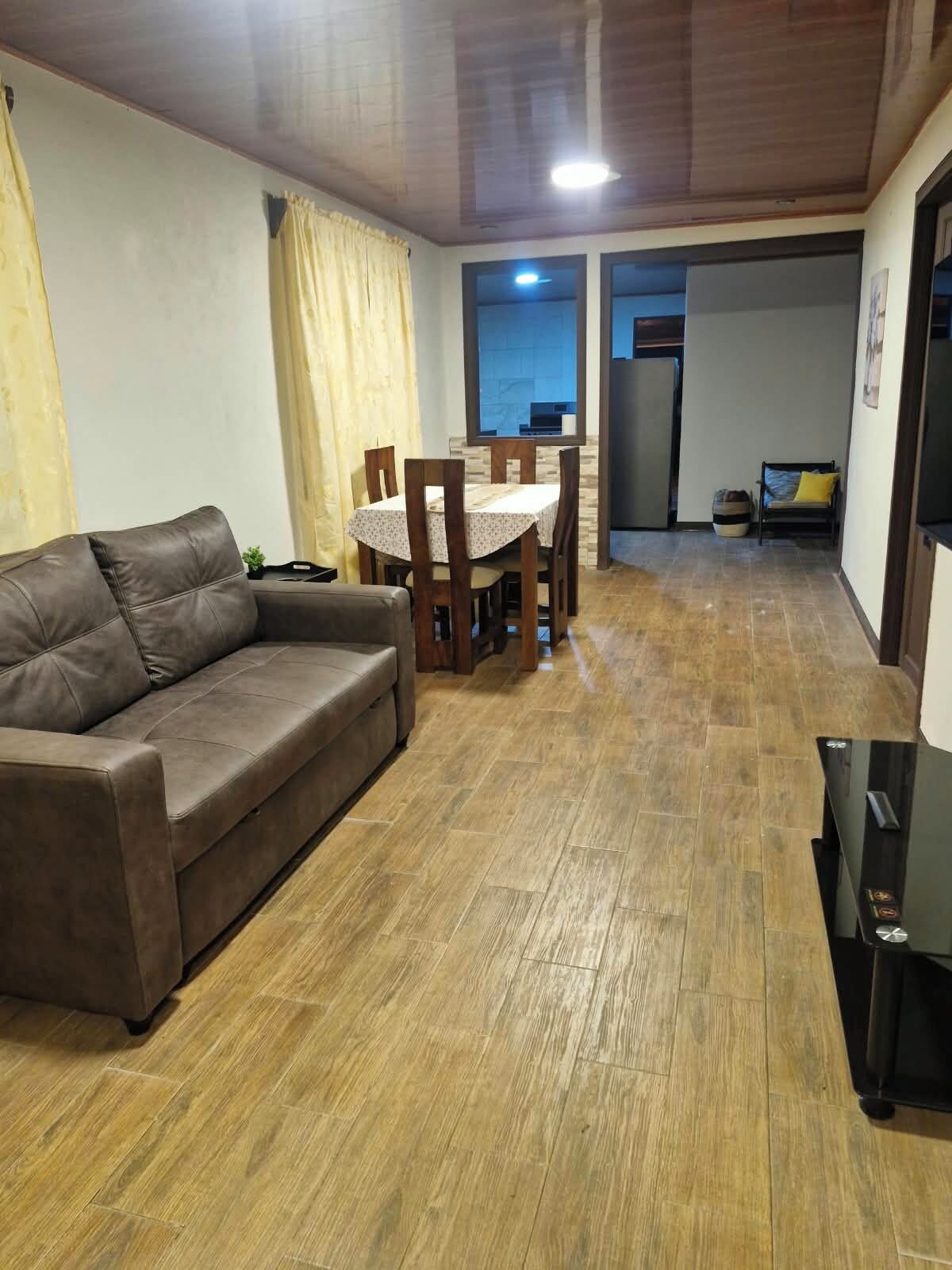 Un salon avec un canapé en cuir marron, une table à manger avec des chaises, et un coin cuisine à l’arrière-plan.