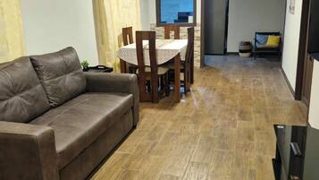 Un salon avec un canapé en cuir marron, une table à manger avec des chaises, et un coin cuisine à l’arrière-plan.