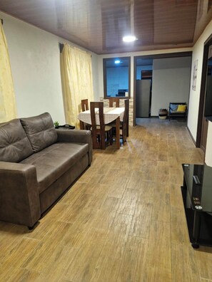 A living room with a brown leather sofa, a dining table with chairs, and a kitchen area in the background.