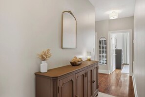 A hallway with a wooden cabinet, a mirror, a vase with dried plants, and a floor lamp. - The Arch House – 4BR – Midtown – Game Loft + Fire Pit (Columbus)