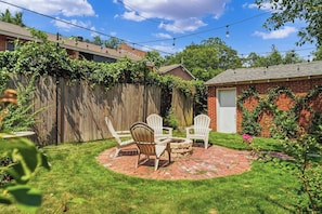 A backyard with a brick fire pit, white Adirondack chairs, a wooden fence, and a brick building. - The Arch House – 4BR – Midtown – Game Loft + Fire Pit (Columbus)