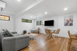 A modern living room with a grey sofa, wooden dining table, and chairs, a flat-screen TV mounted on the wall, and a ceiling fan. - Banksia on the Beach (Gold Coast)