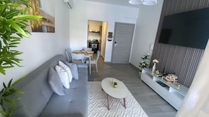 A modern living room with a grey sofa, a small white table, and a TV on the wall. - Resort Style - Pools, Artificial BEACH, Balcony, Fishing Lake, Gulf Club, Gym (Punta Cana)