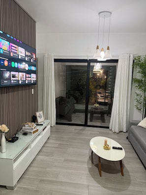 A modern living room with a flat-screen TV displaying various apps, a white console table with decorative items, a round coffee table, and a sliding glass door leading to an outdoor seating area. - Resort Style - Pools, Artificial BEACH, Balcony, Fishing Lake, Gulf Club, Gym (Punta Cana)