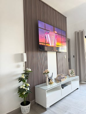 Un salon moderne avec un grand téléviseur à écran plat fixé au mur, qui est recouvert de lambris verticaux, une console blanche agrémentée d’objets décoratifs et une plante en pot.