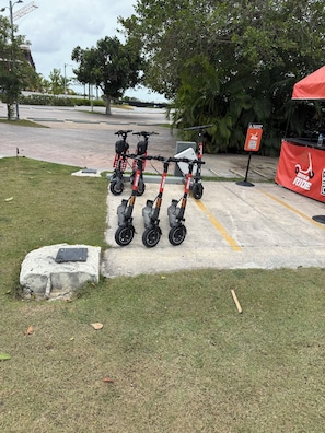 A parking area with several electric scooters lined up, a red banner with 'GRISA RIDE' text, and a sign indicating scooter rental. - Resort Style - Pools, Artificial BEACH, Balcony, Fishing Lake, Gulf Club, Gym (Punta Cana)