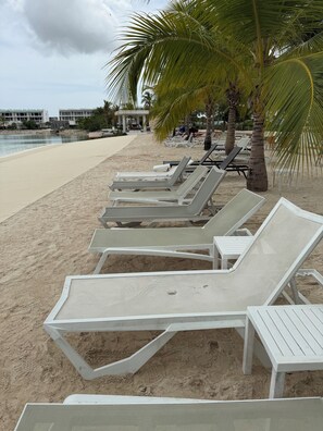 A beach with rows of white sun loungers under palm trees, a sandy shore, and buildings in the background. - Resort Style - Pools, Artificial BEACH, Balcony, Fishing Lake, Gulf Club, Gym (Punta Cana)