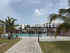 A pool area with lounge chairs, a covered seating area, and a building in the background. - Resort Style - Pools, Artificial BEACH, Balcony, Fishing Lake, Gulf Club, Gym (Punta Cana)
