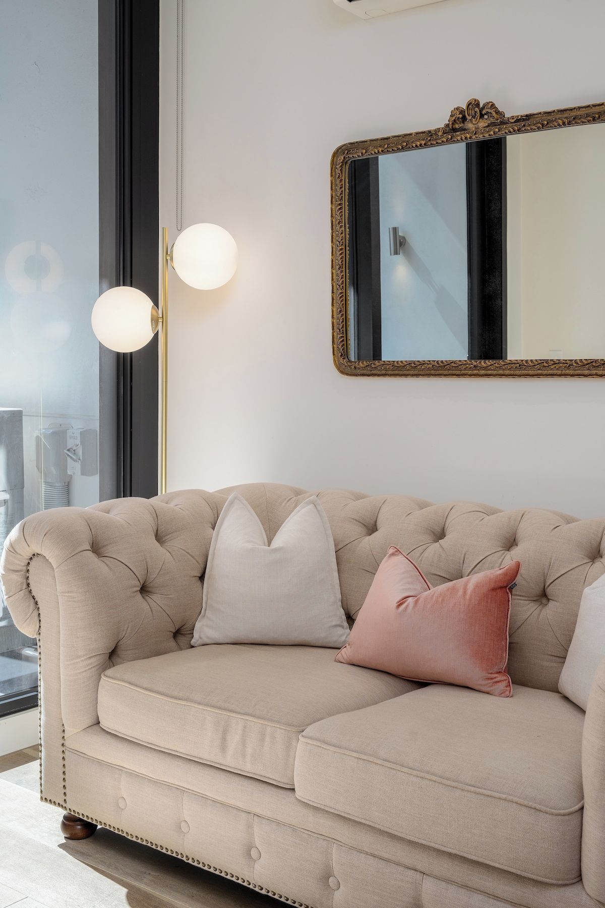 A beige sofa with tufted detailing and a single pink pillow, positioned in front of a large mirror with an ornate gold frame.