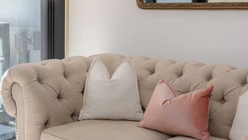 A beige sofa with tufted detailing and a single pink pillow, positioned in front of a large mirror with an ornate gold frame.