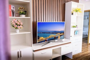 A modern entertainment unit with a flat-screen TV, wooden panel wall, and white shelving with decorative items. - My Khe Ocean View Apartment (Đà Nẵng)