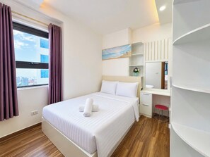 A hotel room with a bed, a desk, a chair, a window with curtains, a painting, and a city view. - My Khe Ocean View Apartment (Đà Nẵng)