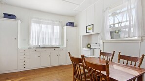 Ein Zimmer mit einem Holzes Esstisch und Stühlen, einem weißen Schrank mit Schubladen, einem Fenster mit leichtem Vorhang und einer an der Wand befestigten Uhr.