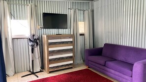 Ein Zimmer mit Fernseher an der Wand, einem Holzschrank, einem violetten Sofa, einem Standventilator und einem roten Teppich.