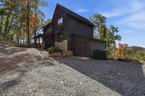A two-story wooden house with a stone foundation, a gravel driveway, and a wooded backdrop. - Black Diamond  Luxury Unwind: Hot Tub, Game Room, Sleeps 14 (Banner Elk)