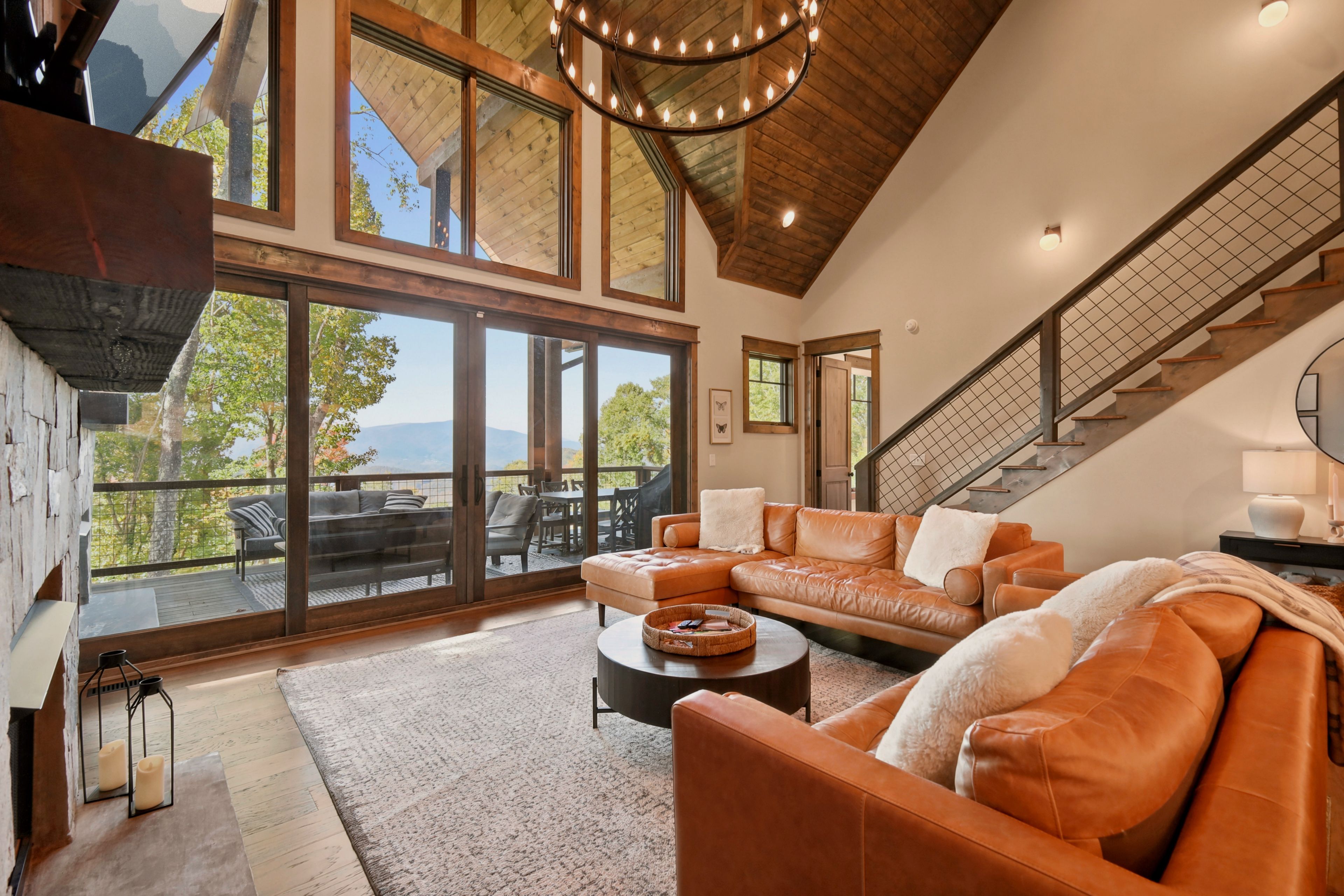 A spacious living room with a large sectional sofa, a coffee table, and a fireplace. There is a staircase leading to an upper level, and large windows offering a view of the outdoors.