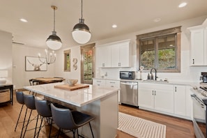 A modern kitchen with a central island, bar stools, a dining area with a wooden table, and a large window providing natural light. - Black Diamond  Luxury Unwind: Hot Tub, Game Room, Sleeps 14 (Banner Elk)