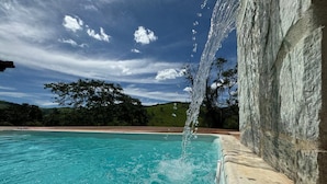 A clear pool with a waterfall feature, surrounded by a stone wall and a view of a green landscape. - Vrbo Property (Moeda)