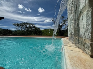 Una piscina cristalina con una cascada, rodeada por un muro de piedra y vistas a un paisaje verde.