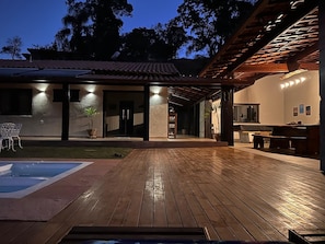 Una terraza de madera con piscina, un porche cubierto y un edificio con tejado de baldosas.