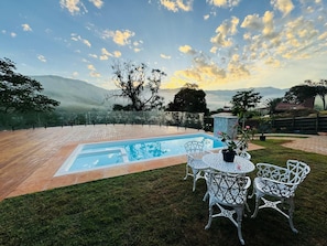 Una piscina con vistas a montañas, rodeada de césped y sillas de metal blanco.