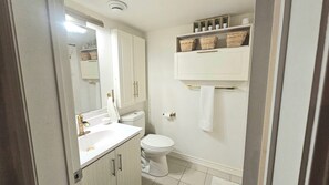 Un baño con un lavamanos blanco, un inodoro y un espejo. Hay cestas en un estante sobre el inodoro.