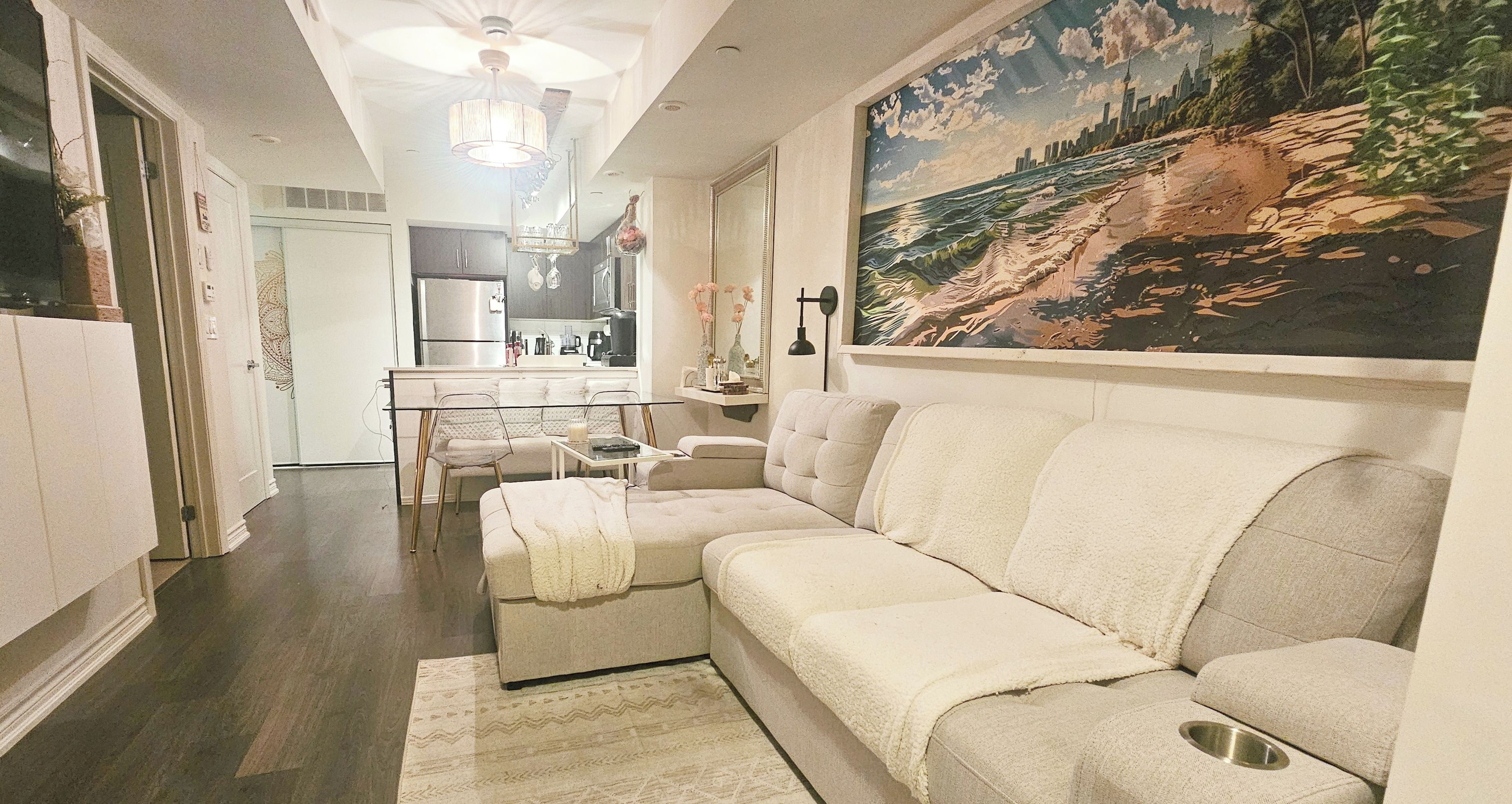 A living room with a sofa, a dining area with a table and chairs, a kitchen in the background, and a large painting on the wall.