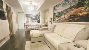 A living room with a sofa, a dining area with a table and chairs, a kitchen in the background, and a large painting on the wall. - Cozy Nook Near the Lake (Toronto)