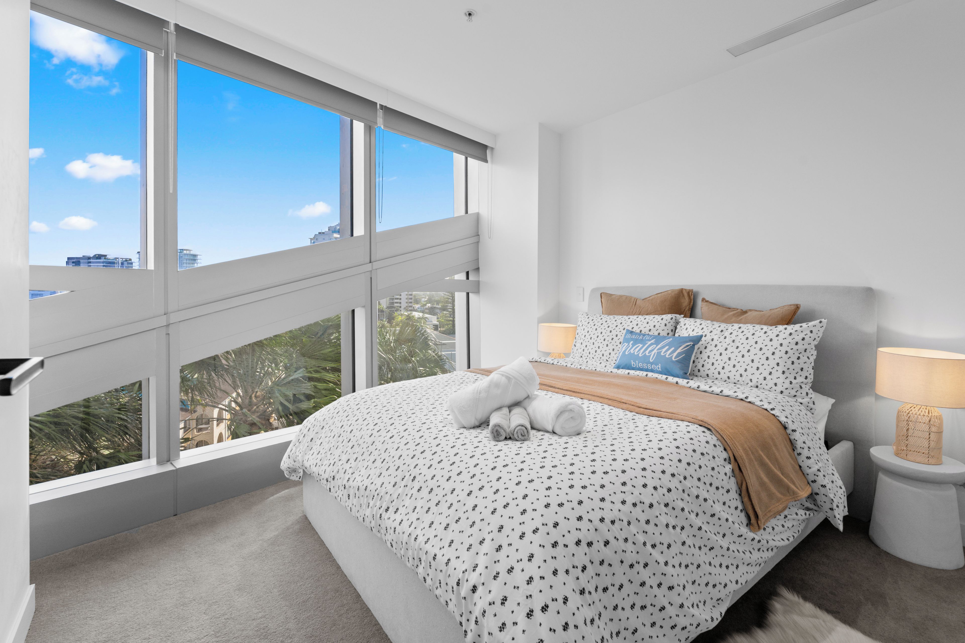A modern bedroom with a large window, a bed with a patterned bedspread, two pillows, a bedside lamp, and a view of the city.