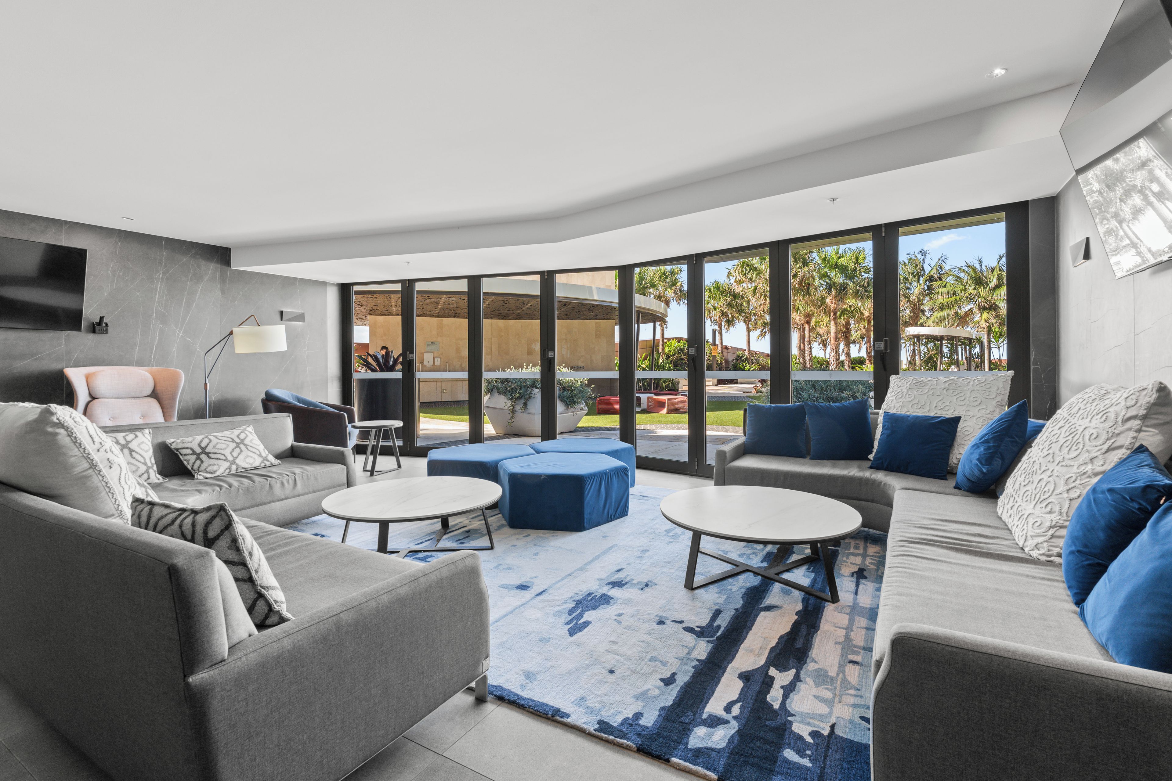 A modern living room with grey sofas, blue cushions, and a large window offering a view of palm trees.