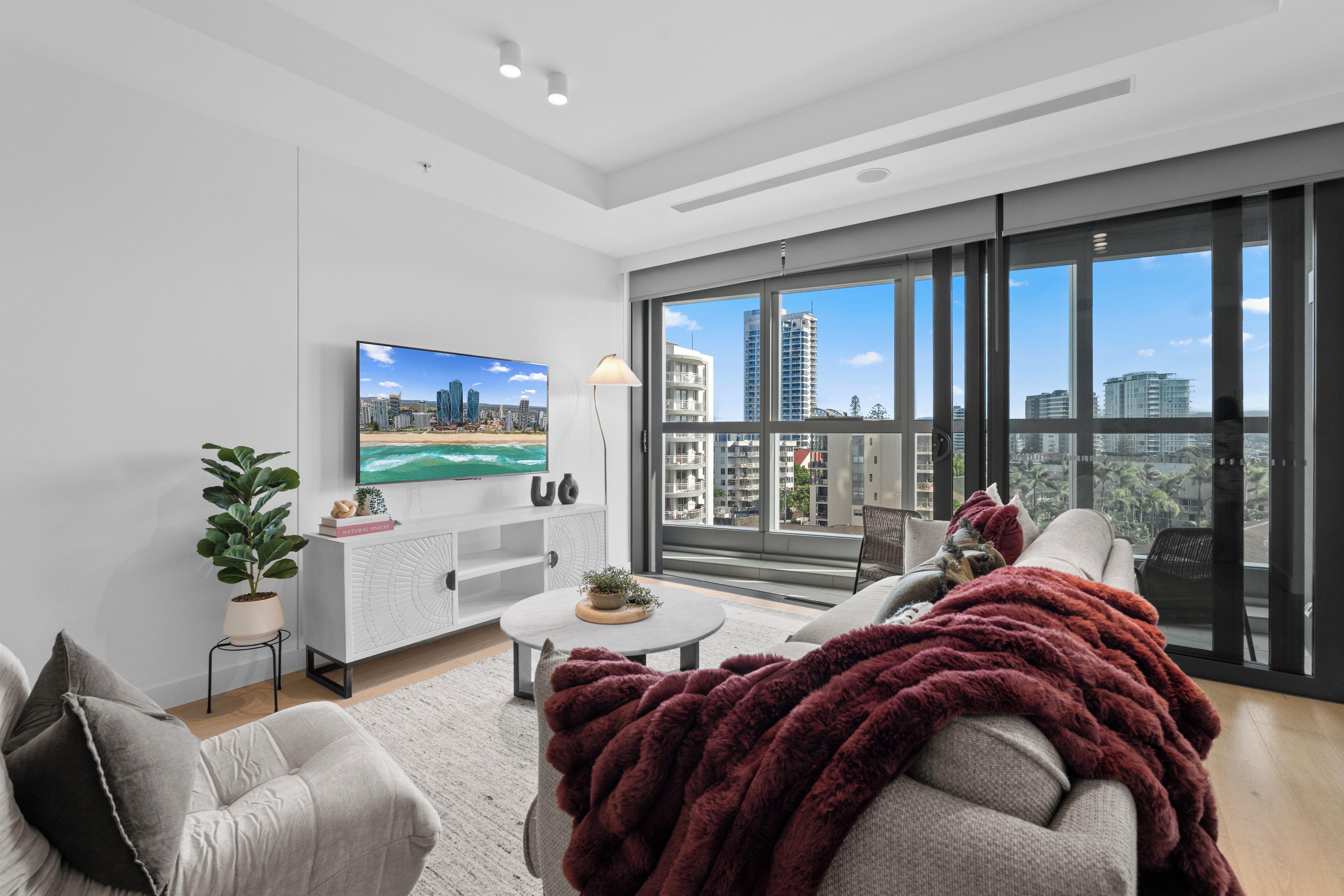 A modern living room with a large TV, comfortable seating, a coffee table, and a view of a cityscape through large windows.
