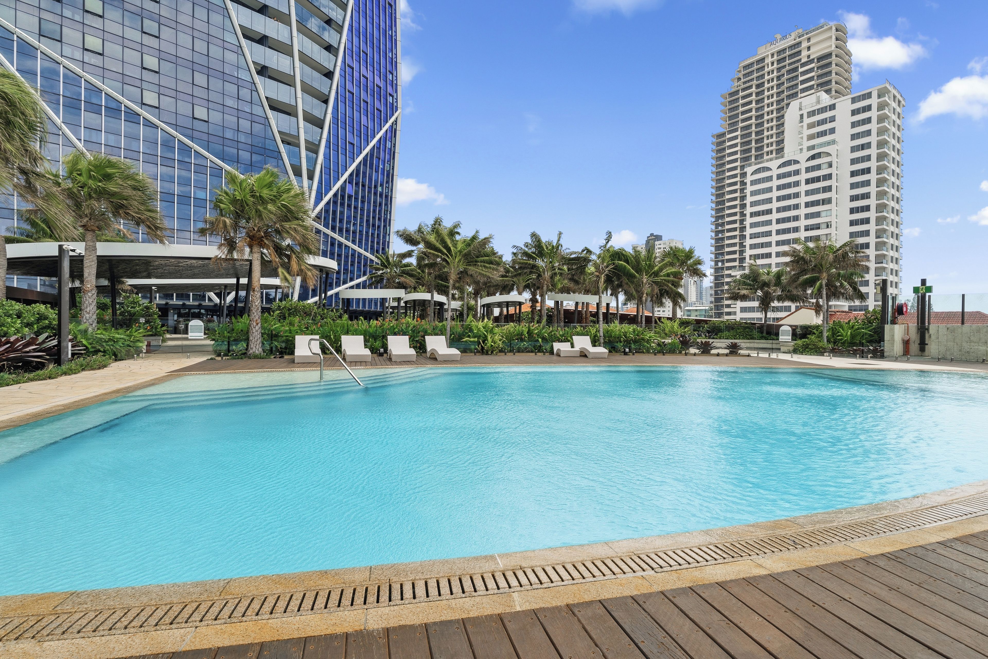 A swimming pool with lounge chairs, palm trees, and modern buildings in the background.