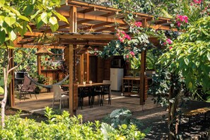 A wooden pergola with a dining area underneath, surrounded by lush greenery and flowering plants. - Venado · 2-BR Family Jungle Home + Pool + AC· Walk to Beach (Cabuya)