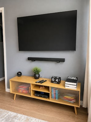 A modern living room with a mounted TV, a wooden entertainment unit with books and a clock, and a potted plant. - 2BD 2BA Near UH & Downtown, balcony + free parking (Houston)