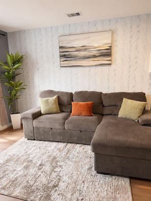 A living room with a sectional sofa, a large abstract painting, a potted plant, and a textured rug. - 2BD 2BA Near UH & Downtown, balcony + free parking (Houston)