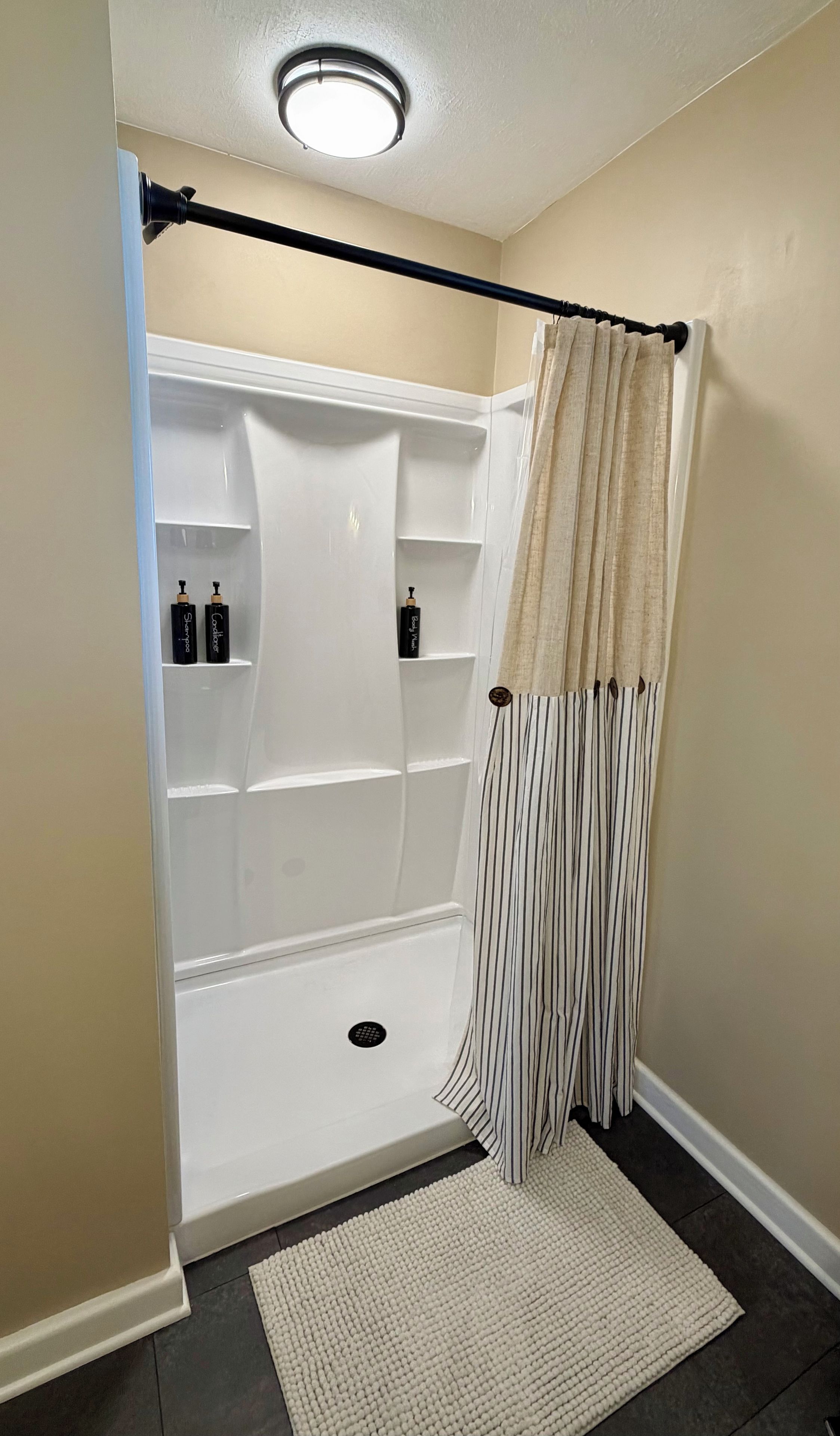 Une douche à l’italienne avec un intérieur blanc, un rideau beige et deux bouteilles posées sur les étagères.