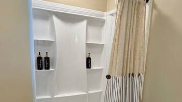 Une douche à l’italienne avec un intérieur blanc, un rideau beige et deux bouteilles posées sur les étagères.