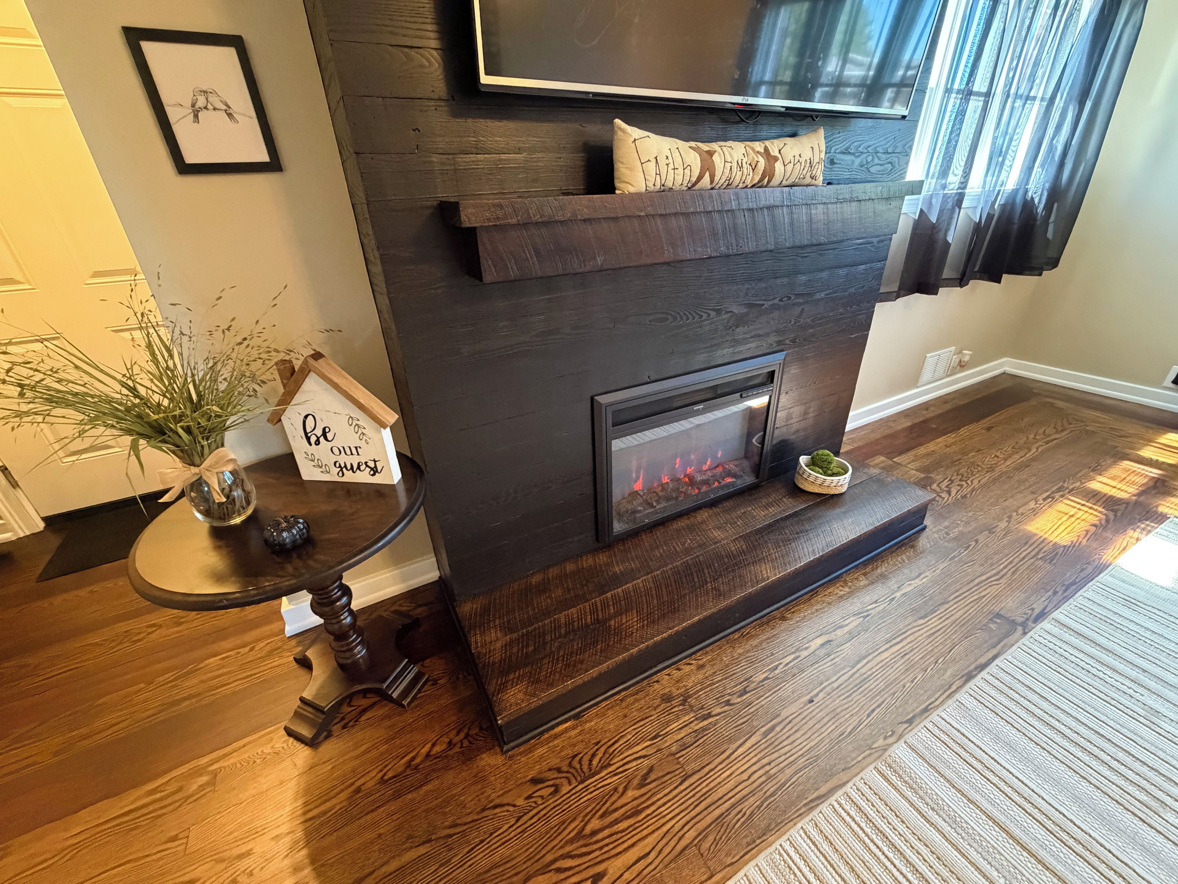Un salon moderne avec une cheminée en bois foncé, un téléviseur à écran plat fixé au-dessus, et une petite table d’appoint agrémentée d’une plante et d’un panneau.