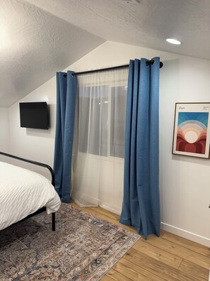 Une chambre à coucher avec un lit, un téléviseur fixé au mur, une fenêtre avec des rideaux légers et un tableau encadré accroché au mur.