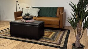 Een woonkamer met een bruine leren bank, een zwart poufje, een vloerlamp, een groene plaid, een geweven vloerkleed en een potplant.