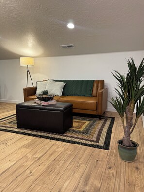Un salon avec un canapé en cuir marron, un pouf noir, une lampe sur pied, un plaid vert, un tapis tissé et une plante en pot.