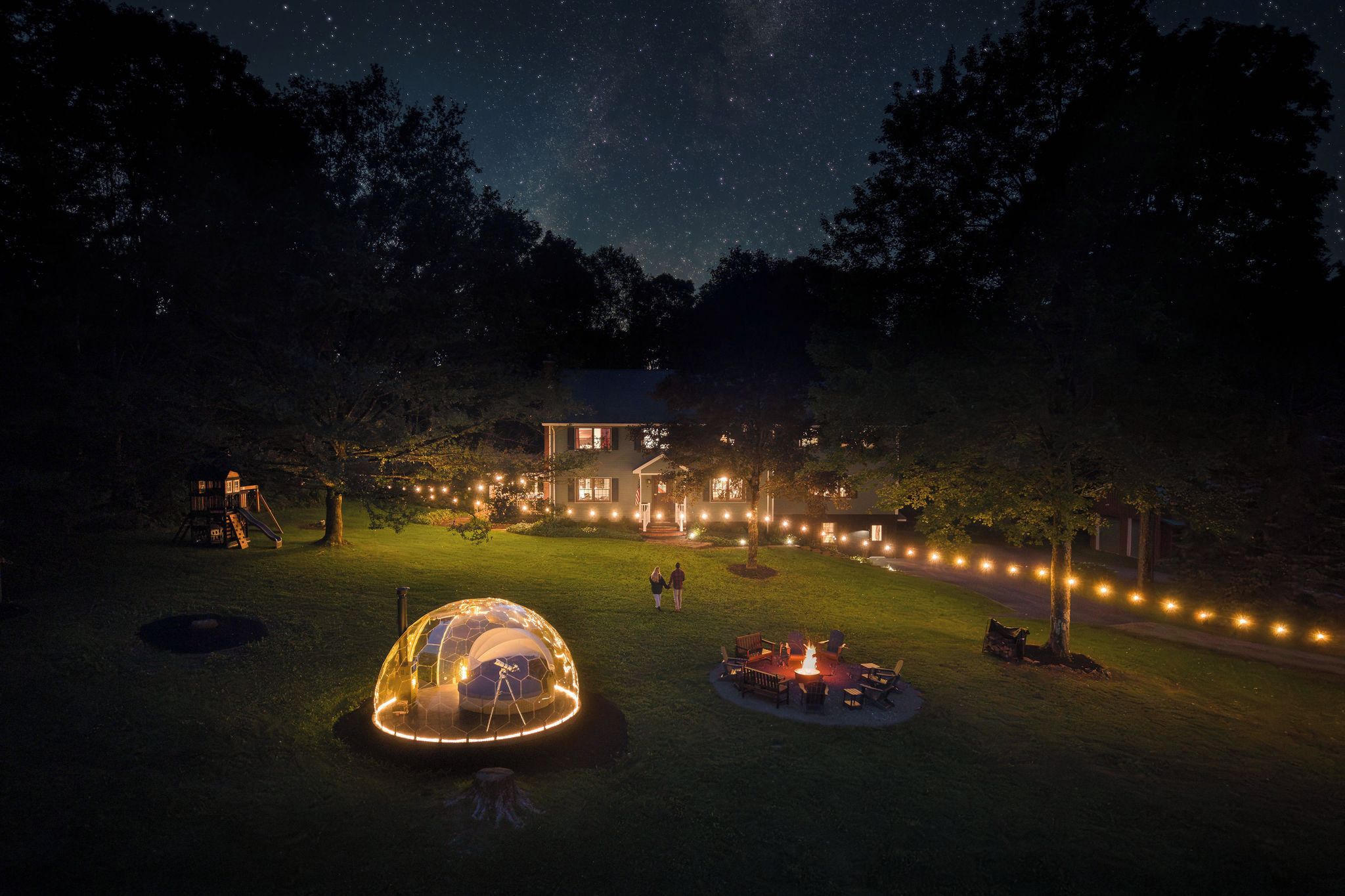 A house with a dome-shaped structure in the foreground, a fire pit, and a string of lights along the pathway.